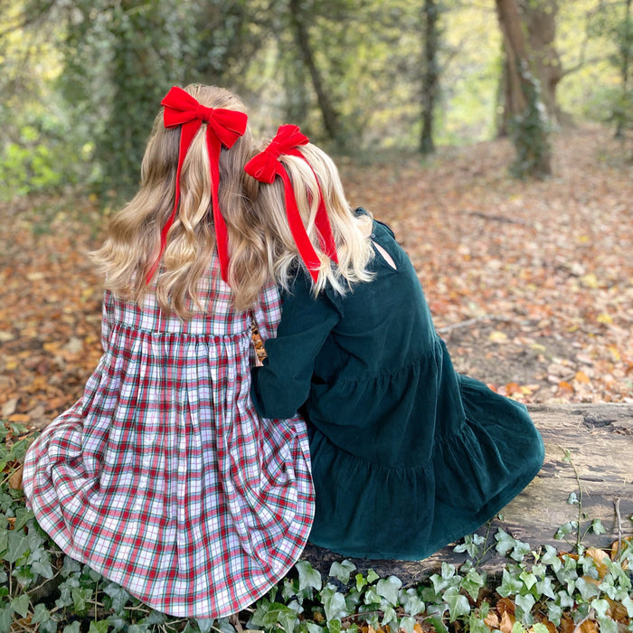 Two children with red velvet bows in their hair sitting on a log in a forest. Girls Wearing a Tartan Dress and a green dress for Christmas with matching red hair bows