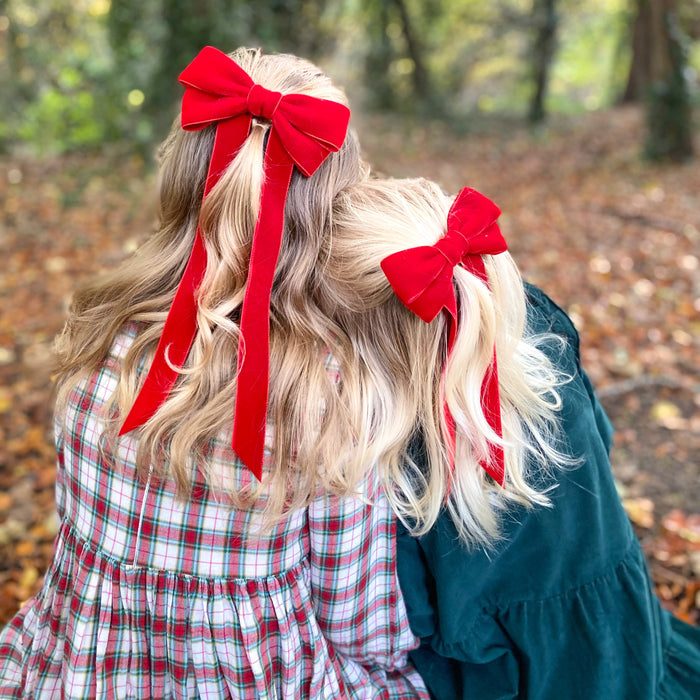 Two children with matching red velvet bows in their hair sitting in a forest with fallen leaves on the ground. Sister sChristmas outfit matching hair bows
