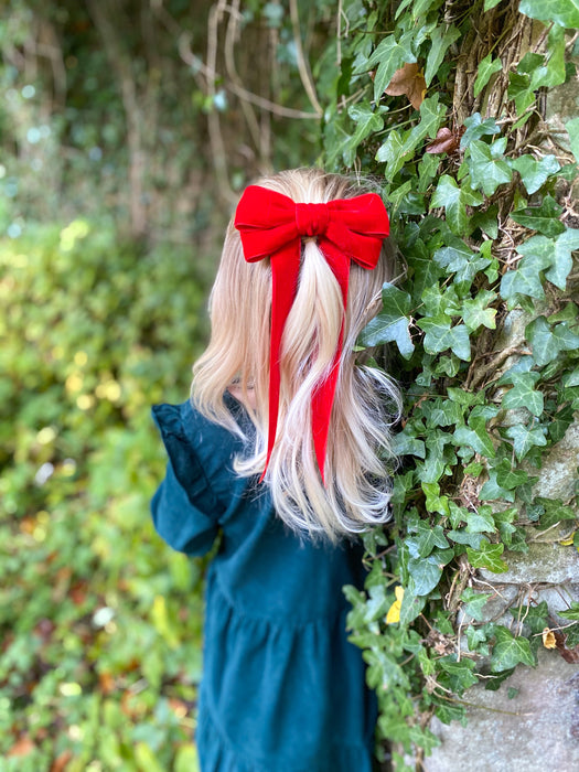 Childs Little Love red velvet bow hair clip worn by little girl in forest with long blonde hair and a green dress , leaning against an ivy wall, a festive scene