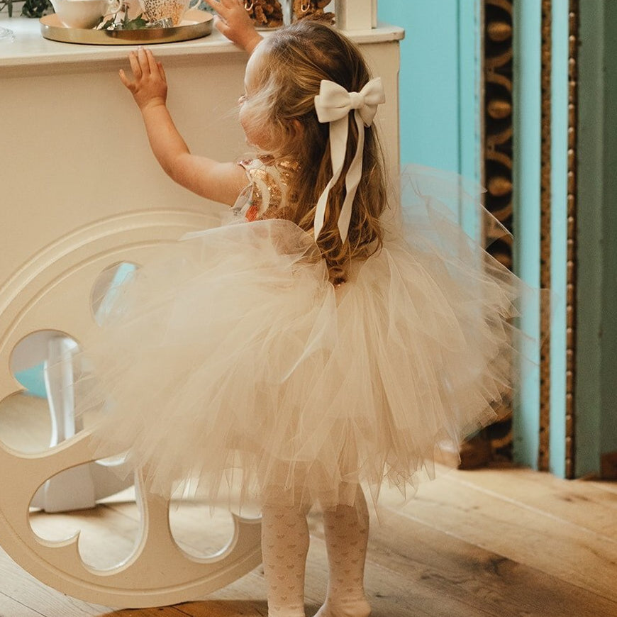 A toddler flower girl wearing a fluffy cream tulle tutu dress with a matching Velvet bow in her hair, standing in a room and reaching toward a white cabinet at a wedding