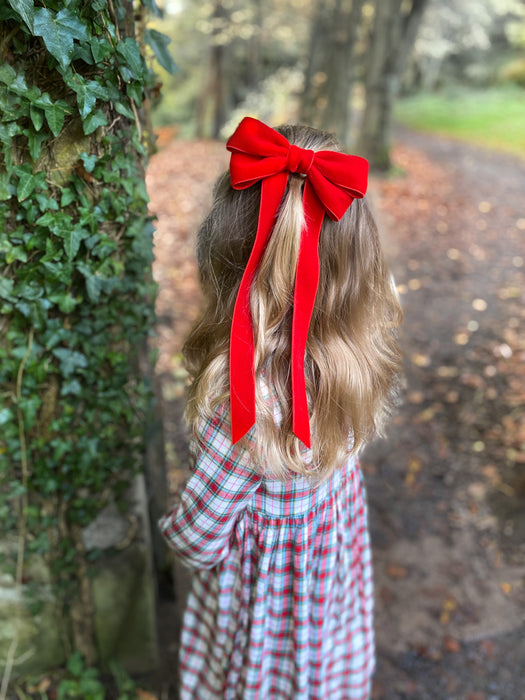 young girl with red velvet bow in their hair standing by tree in forest. Girl Wearing a Tartan Dress for Christmas with matching red hair bow